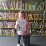 Powiększ zdjęcie W Bibliotece Hania laureatka konkursu trzymająca dyplom obok stoi torba z nagrodą 