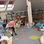 Powiększ zdjęcie Dzieci siedzą na podłodze w bibliotece chłopiec wybiera kolorowy liść leżący na podłodze