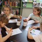 Powiększ zdjęcie Dzieci siedzą przy stolikach i kolorują dinozaura