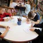 Powiększ zdjęcie Dzieci siedzą przy stolikach i kolorują dinozaura