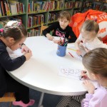 Powiększ zdjęcie Dziewczynki siedzą przy stoliku i kolorują dinozaura